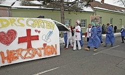 Eine Gruppe von Menschen in blauen Kitteln geht auf einer Straße mit einem Banner, das 'Drs. Carten Herzsation' mit einem Herz und einem roten Kreuz zeigt.