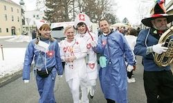 Fünf Personen in blauen Kostümen und Masken gehen auf einer Straße, zwei halten sich an den Händen und einer spielt ein Blasinstrument.