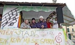 Eine Gruppe von Menschen sitzt in einer dekorierten Kabine mit musikalischen Noten an der Wand, einige tragen Winterkleidung, und ein Banner mit deutschem Text befindet sich über ihnen.