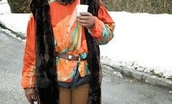 Eine Person steht auf einer verschneiten Straße und hält einen Becher. Sie trägt ein farbenfrohes oranges Shirt und einen Pelzmantel.
