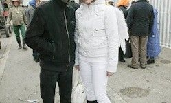 Ein Mann und eine Frau stehen zusammen auf einem Bürgersteig, der Mann in einer schwarzen Jacke und die Frau in einem weißen Skianzug mit Engelsflügeln. Hinter ihnen laufen mehrere Leute auf dem Bürgersteig.