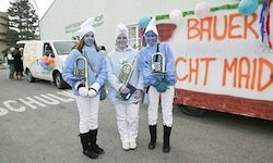 Drei Personen in blauen Kostümen und mit Musikinstrumenten stehen vor einem dekorierten Wagen. Der Wagen hat einen weißen Hintergrund mit dem Wort 'BAUER' und ein blaues Banner mit dem Wort 'CHCHT MAID'.