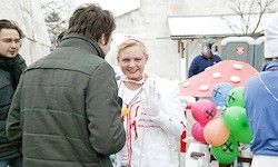 Eine Frau in einem weißen Mantel, der Luftballons hält, spricht mit einem Mann in einer grauen Jacke. Dahinter stehen andere in der Nähe einer pilzförmigen Struktur.