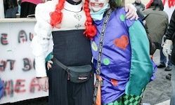 Zwei Menschen in Kostümen posieren für ein Foto. Einer trägt eine rote Perücke und ein schwarzes Outfit. Der andere trägt ein farbenfrohes Clownkostüm mit einer Bauchtasche. Sie stehen vor einem Banner.