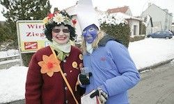Zwei Frauen in Kostümen stehen im Schnee. Eine trägt eine blaue Gesichtsbemalung, die andere ein blumenverziertes Kostüm. Ein Haus und ein Auto sind im Hintergrund.