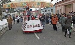 Ein festlicher Wagen fährt auf einer verschneiten Straße, viele Menschen gehen um ihn herum. Der Wagen trägt ein Schild mit der Aufschrift 'Es Danke'.
