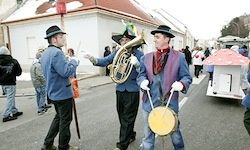 Drei Musiker in blauen Anzügen und Hüten spielen Instrumente bei einer Parade auf einer Straße. Dahinter sind eine Gruppe von Menschen und eine rosa Struktur zu sehen.