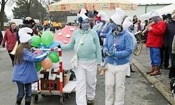 Eine Gruppe von Menschen in Kostümen, einschließlich mit blauer Gesichtsbemalung, geht mit einem Wagen voller Luftballons. Ein Kind mit einem weißen Hut hält sich am Wagen fest. Im Hintergrund spielen Menschen in Kostümen Instrumente.
