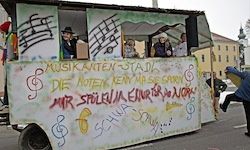 Ein dekorierter Wagen mit Graffiti zeigt eine Nachricht, die 'Musicanten-Stand' und 'Die Noten Keny-Hap Se Garrn' lautet. Eine Person in einer blauen Jacke läuft auf der Straße.