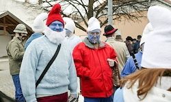 Menschen in Kostümen und Gesichtsbemalung gehen auf einer Straße, einer Mann in einem roten Mantel und einer in einem blauen Pullover. Sie sind von anderen in weißen Hüten umgeben, der Hintergrund ist verschwommen.