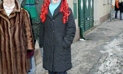 Zwei Frauen mit leuchtend roten Haaren, eine in einem Pelzmantel und die andere in einer schwarzen Daunenjacke, gehen auf einer verschneiten Straße. Hinter ihnen steht eine Person in einem grünen Mantel in der Nähe eines Gebäudes.