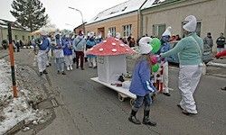 Eine Gruppe von Menschen in weißen Kostümen, Mützen und Schuhen läuft bei einer Parade. Einer schiebt einen Wagen mit einem pilzförmigen Dach. Im Hintergrund sind Gebäude und Bäume zu sehen.