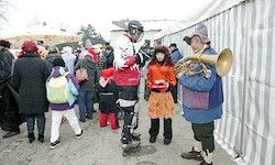 Eine Gruppe von Menschen in verschiedenen Kostümen steht in einer Reihe. Ein Mann spielt Posaune, während eine Frau auf Rollerskates ein Kind hält. Im Hintergrund sind Bäume verschwommen.