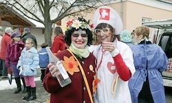 Zwei Menschen in Kostümen stehen zusammen; eine in einem Blumenkleid und der andere in einer Krankenschwesteruniform, posieren für ein Foto. Dahinter stehen mehrere Personen, einige in Winterkleidung. Ein Baum und ein Gebäude sind im Hintergrund zu sehen.