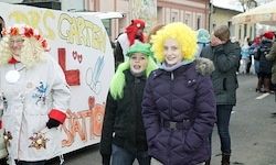Drei Menschen in Kostümen stehen auf der Straße, einer trägt einen grünen Hut. Ein Banner ist hinter ihnen. Einer von ihnen hat einen Gürtel. Eine weitere Person trägt einen roten Hut.