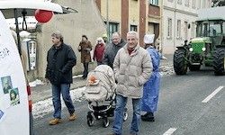 Eine Gruppe von Menschen geht auf einer Straße. Einige sind warm in Winterkleidung gekleidet. Ein Mann schiebt einen Kinderwagen mit einem Baby. Ein Traktor ist am Straßenrand geparkt.