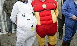 Zwei Personen stehen nebeneinander, einer in einem roten und gelben Iron-Man-Kostüm und der andere in einer weißen Schutzkleidung. Hinter ihnen beobachten drei weitere Personen in blauer Schutzkleidung.