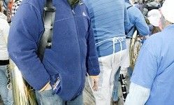 Ein Mann in einer blauen Jacke und weißen Hosen steht unter anderen, möglicherweise bei einer Marschveranstaltung, und trägt ein Tuba.