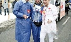 Drei Menschen in medizinischen Kostümen posieren für ein Foto auf einer verschneiten Straße. Einer trägt einen blauen Kittel, ein anderer einen weißen Kittel, und ein Krankenwagen ist im Hintergrund zu sehen.
