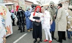 Zwei Menschen in weißen Kostümen mit roten Perücken posieren für ein Foto. Sie sind von Menschen in verschiedenen Kostümen umgeben, einige in blauen Laborkittel und andere in Winterkleidung, die auf einer Straße stehen.