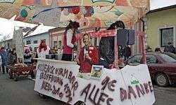 Ein Straßenwagen mit Personen darin steht auf der Straße. Es gibt Luftballons und Bilder, die befestigt sind. Eine Person in Rot steht davor. Mehrere Menschen und ein Auto sind dahinter. Gebäude befinden sich an der Seite.