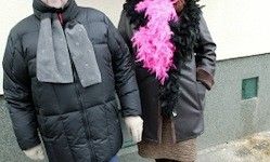 Zwei Personen in Winterkleidung, eine mit einem rosa Federboa, stehen im Freien in der Nähe einer Wand.