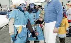 Drei Menschen in blauen Kostümen mit Gesichtsbemalung, darunter ein Mann in einem blauen Jackett, posieren für ein Foto auf einer verschneiten Straße. Hinter ihnen ist ein Auto geparkt, und mehrere Personen in verschiedenen Kostümen sind anwesend.