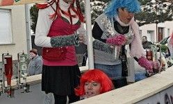 Zwei Frauen mit leuchtend roten Perücken stehen hinter einem Tresen, eine hält ein Glas und die andere greift nach etwas, während eine Frau mit einer roten Perücke hinter dem Tresen hervorschaut.