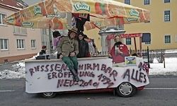 Ein kleiner Wagen mit einem gelben Sonnenschirm hat Graffiti an der Seite, die 'Riessenheim Eroffnung Hirm Bar' und 'Neue 2013' lesen. Ein Mann sitzt auf der Theke, hinter ihm steht eine Frau.