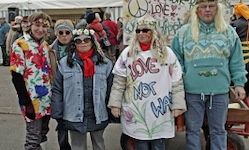 Fünf Menschen, gekleidet in farbenfrohe Kleidung und Winteroutfits, stehen gemeinsam im Freien. Eine Frau trägt ein Shirt mit der Aufschrift 'LIEBE NICHT KRIEG'.
