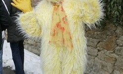 Eine Person in einem gelben pelzigen Kostüm mit roten Designs steht draußen in der Nähe einer Steinmauer und Schnee. Hinter ihr steht eine weitere Person in einem schwarzen Mantel und gelben Handschuhen.