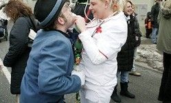 Ein Mann in einem blauen Anzug und eine Frau in einem weißen Outfit mit rotem Rand küssen sich auf der Straße. Dahinter stehen mehrere Menschen in Winterkleidung und beobachten.