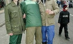Drei Personen stehen draußen; zwei Männer in Jacken, einer mit einem Getränk, und ein Junge in Schwarz hinter ihnen. Der Hintergrund zeigt eine verschwommene Straße mit Menschen.
