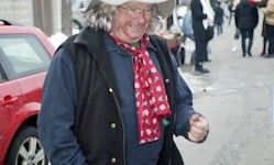 Ein älterer Mann mit weißen Haaren, in einem blauen Shirt, schwarzem Weste, rotem Schal und Hut, steht auf einem Bürgersteig neben einem roten Auto. Dahinter gehen mehrere Leute in die entgegengesetzte Richtung.