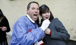 Ein Mann in einem blauen Kittel und eine Frau in einem grauen Mantel stehen eng beieinander, lächeln und lachen. Der Mann trägt weiße Handschuhe, während die Frau ein rotes Langarmshirt unter ihrem Mantel trägt. Eine weitere Frau steht in der Nähe in einem schwarzen Oberteil.