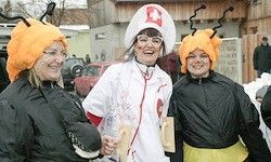 Drei Frauen in Kostümen stehen zusammen. Zwei tragen schwarze Regenmäntel und gelbe Bienenhüte. Eine ist als Krankenschwester mit einem roten Kreuz gekleidet. Sie lächeln vor einem Gebäude.