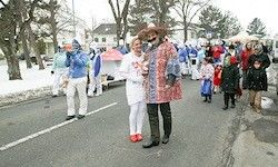 Ein Umzug mit Menschen in Kostümen auf einer verschneiten Straße. Eine Person in einem bunten Mantel und eine andere in einem weißen Outfit stehen in der Mitte. Schnee bedeckt den Boden und die Bäume.