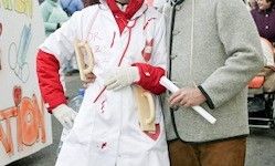 Eine Person in einem blutbefleckten Laborkittel und Handschuhen steht neben einer weiteren in einem grauen Mantel, möglicherweise bei einem Festival oder einer Parade, mit einem rot-weißen Banner im Hintergrund.