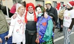 Drei Menschen in Kostümen posieren für ein Foto auf einer überfüllten Straße. Die Frau auf der linken Seite trägt eine Krankenschwester-Kostüm. Der Mann in der Mitte hat ein Clown-Gesicht und rote Haare. Die Frau auf der rechten Seite trägt ein buntes Kostüm.