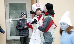 Eine Gruppe von Menschen in verschiedenen Kostümen steht draußen. Eine Frau in einem weißen Mantel und ein Mann in einer grauen Jacke küssen sich. Eine weitere Person hält eine Flasche.
