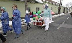 Eine Gruppe von Menschen in blauen Regenmänteln und Mützen zieht in einer Parade auf einer verschneiten Straße. Eine Person trägt eine weiße Maske und hält einen Wagen mit bunten Luftballons.