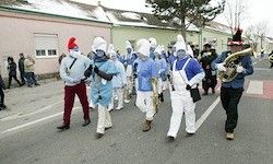 Eine Gruppe von Menschen in blauen Kostümen und Mützen marschiert auf der Straße und spielt Musikinstrumente. Sie werden von Schaulustigen am Bürgersteig begleitet.