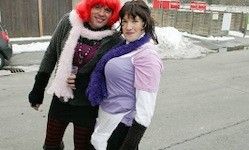 Zwei Personen in farbenfroher Winterkleidung stehen auf einer verschneiten Straße. Eine hält ein Glas, und beide tragen Schals. Ein Gebäude und ein Auto sind im Hintergrund.