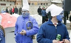 Zwei Männer in blauen Kostümen mit Gesichtsbemalung und Hüten stehen in einem schneebedeckten Bereich. Einer hält ein Musikinstrument. Hinter ihnen stehen und gehen Leute. In der Ferne befindet sich ein großes rosa Objekt.