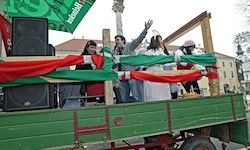 Eine Gruppe von Menschen auf einem grünen Wagen, der mit roten und grünen Bändern verziert ist. Einige winken, während andere lächeln. Ein Lautsprecher ist am Wagen befestigt.