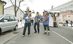 Drei Männer in Kostümen spielen Blech- und Akkordeoninstrumente auf einer Straße mit geparkten Autos und vorbeigehenden Menschen. Die Umgebung umfasst Gebäude und einen schneebedeckten Boden.