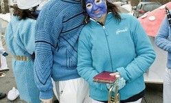 Eine Gruppe von Menschen in blauen Kostümen, von denen eine Person eine blaue Gesichtsmaske hat, ein rotes Objekt hält und neben einem Auto steht.