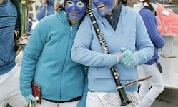 Zwei Personen, in Blau gekleidet und mit blau bemalten Gesichtern, stehen zusammen. Einer hält eine Klarinette und der andere ein Buch. Sie nehmen wahrscheinlich an einer Parade oder Veranstaltung teil.