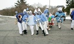 Eine Gruppe von Menschen in blauen Kostümen und weißen Hüten marschiert mit Musikinstrumenten spielend die Straße hinunter, mit verschneiten Bäumen im Hintergrund.