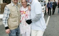 Drei Menschen in Halloween-Kostümen stehen zusammen auf einer Straße, mit Fußgängern und Fahrzeugen im Hintergrund.
